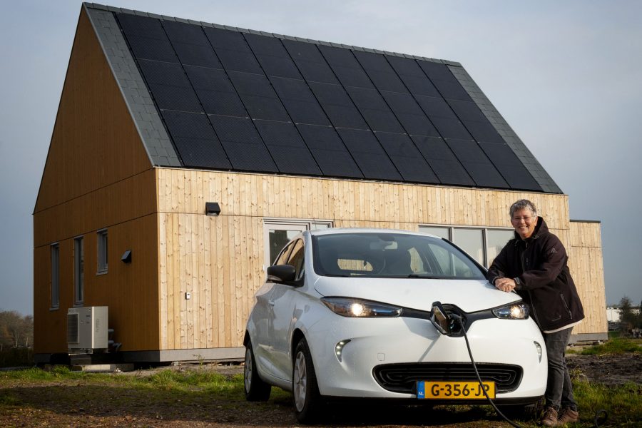 Een vrouw staat naast een elektrische auto voor een huis met zonnepanelen.