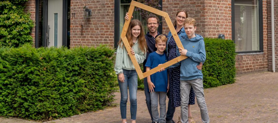 Een gezin poseert voor een huis met een houten frame in de hand.